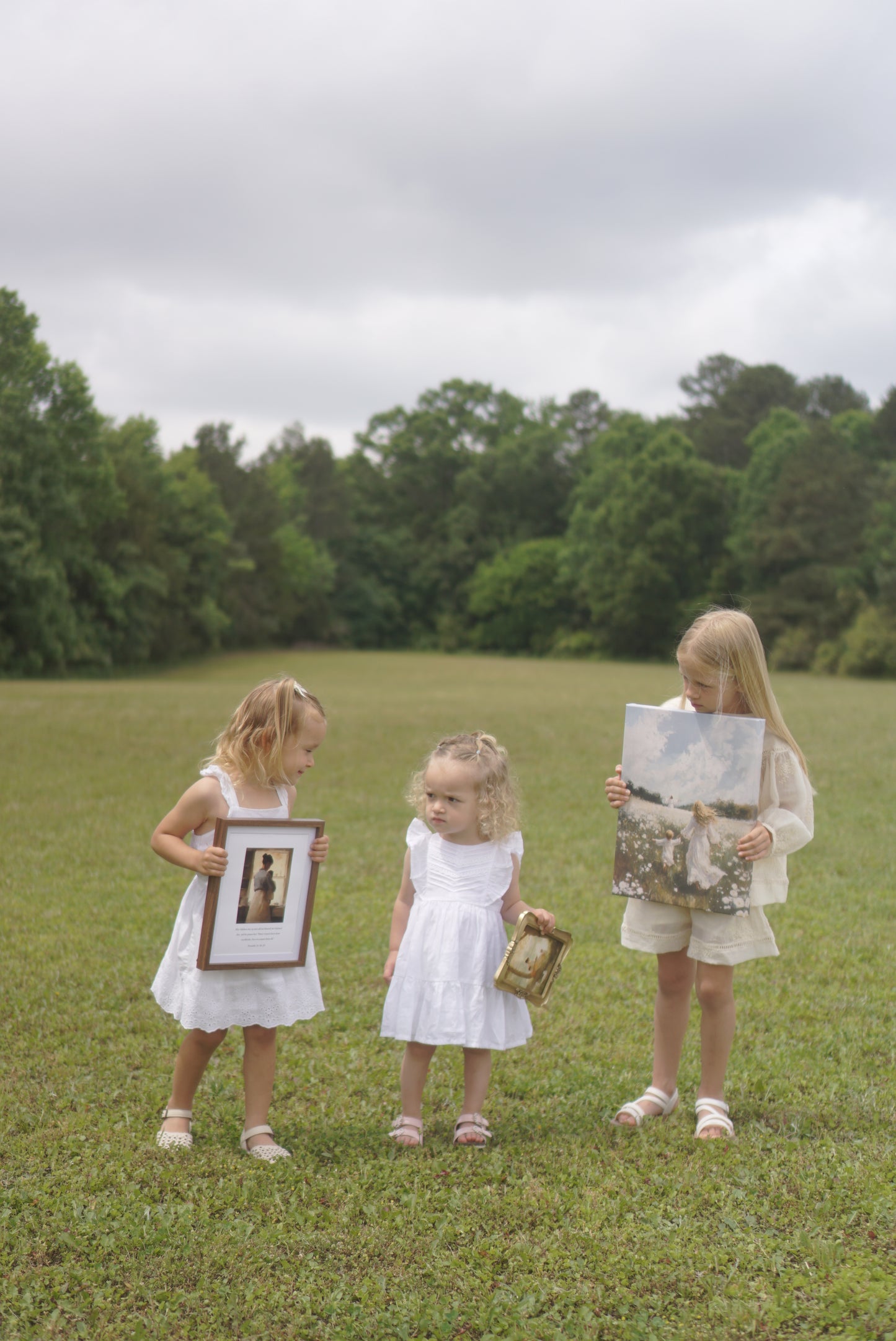Three young girls in white dresses holding framed pictures on a grassy field. Following Him Canvas modern Christian wall art by Christian artist, walking with Jesus in faith.