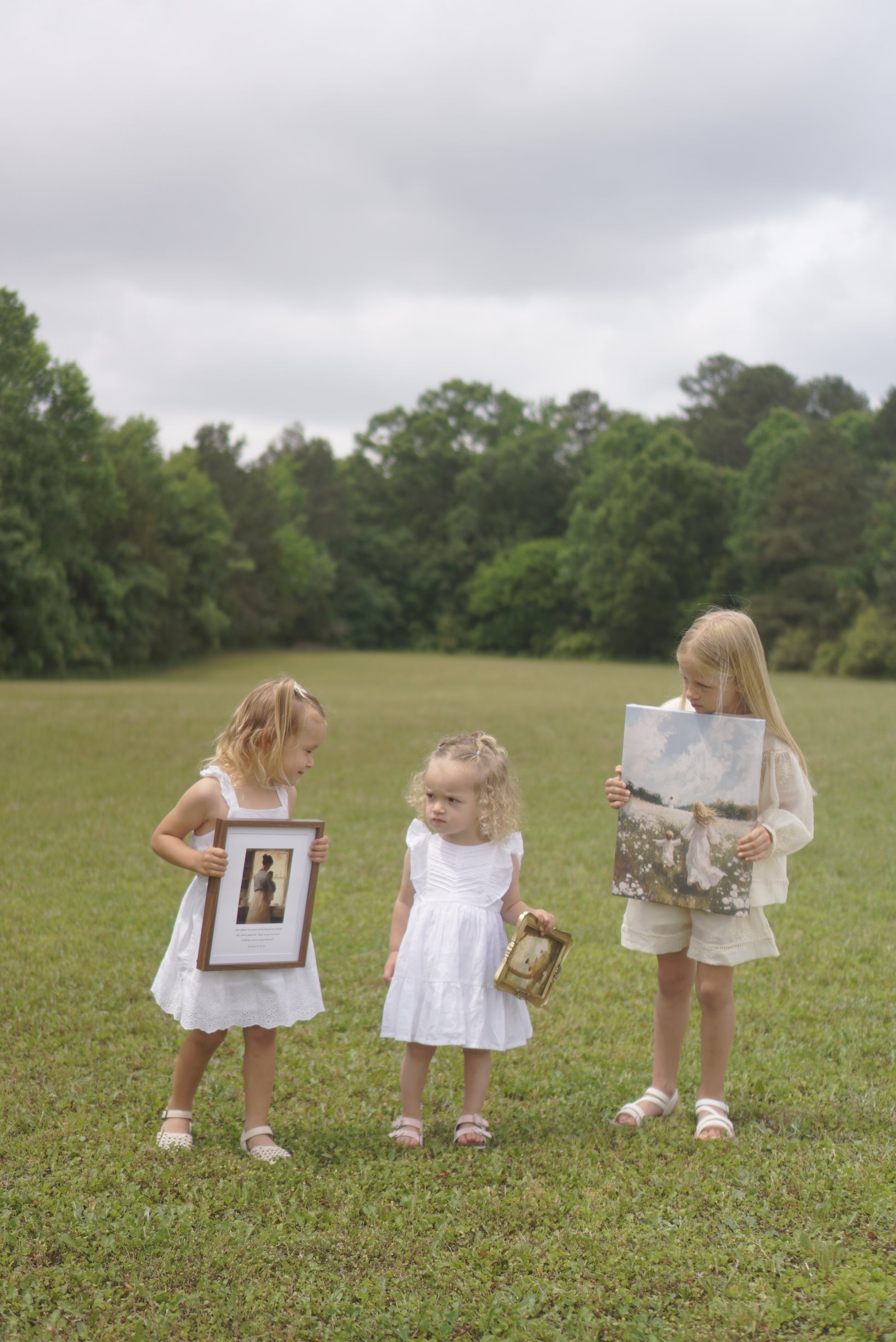 Three young girls in white dresses holding framed pictures on a grassy field. Following Him Canvas modern Christian wall art by Christian artist, walking with Jesus in faith.