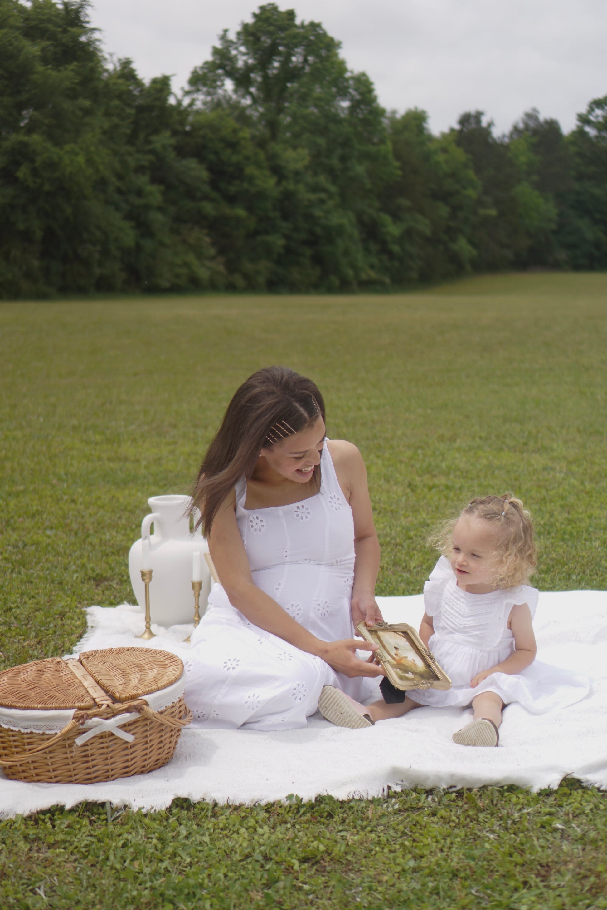 Woman and child in white dresses sitting on a picnic blanket in a grassy field.By the Pond Christian wall art modern by Christian artist capturing peace in God’s presence.