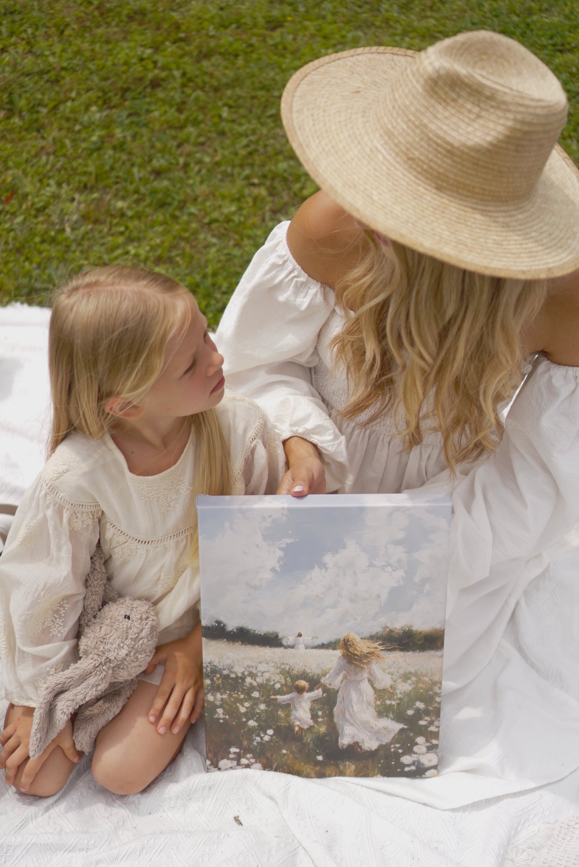 Woman and child sitting on a blanket with a painting of a field and child holding a teddy bear. Following Him Canvas modern Christian wall art by Christian artist, walking with Jesus in faith.